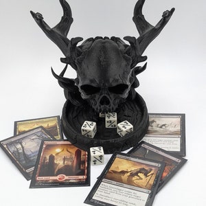 May include: Black skull-shaped dice tower with antlers, surrounded by five black-bordered Magic: The Gathering cards and five white dice. The cards include "Gravedigger", "Bloodhunter Bat", "Mountain", "Basic Land", and "Supernatural Encounter".
