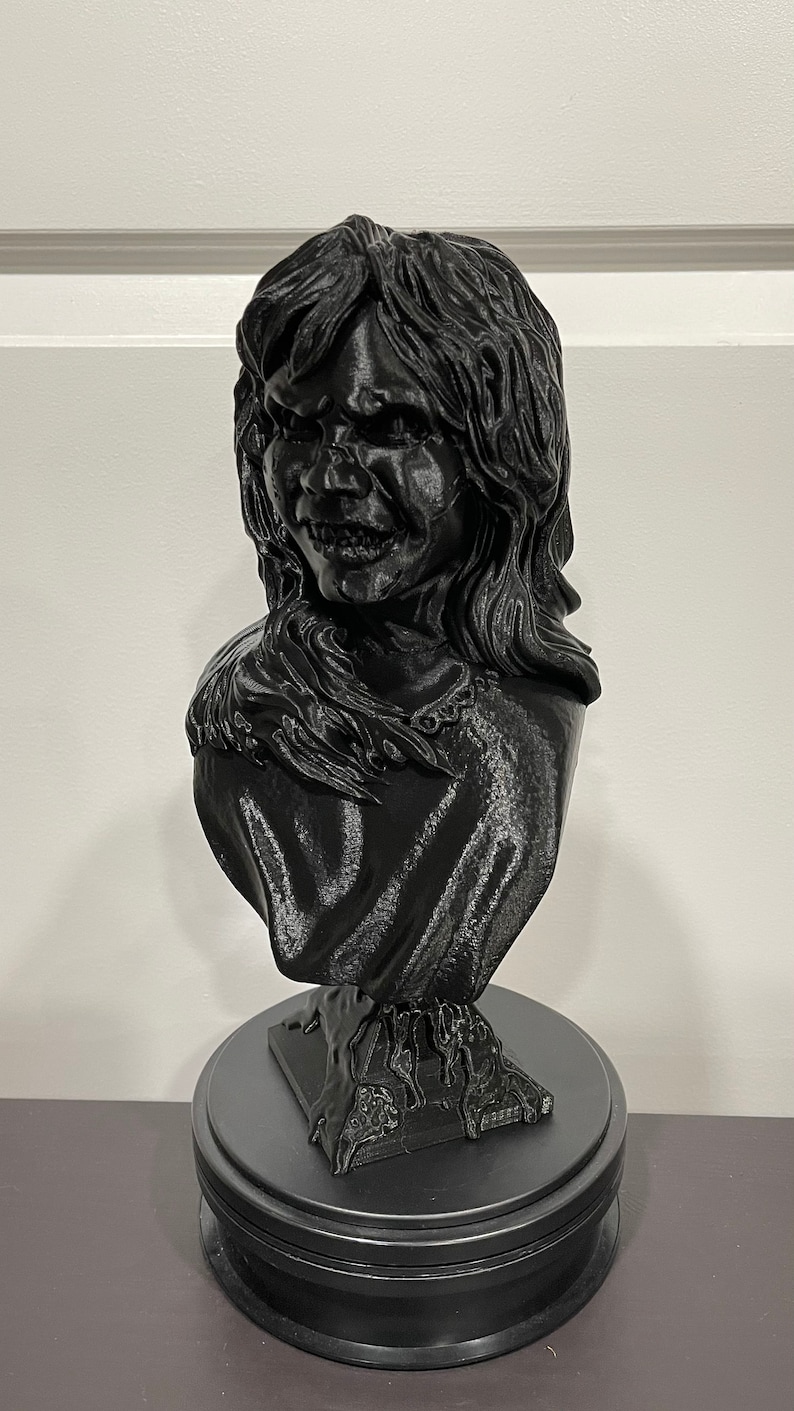 May include: A black 3D printed bust of a girl with long hair and a creepy smile. The bust is on a black circular base.