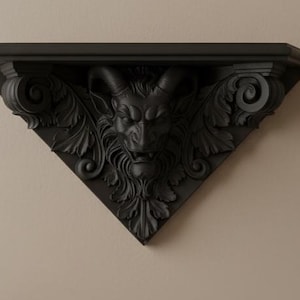 May include: Black, triangular wall shelf with a detailed, carved design. The shelf features a central demonic face with horns, surrounded by ornate leaves and scrollwork. The shelf is mounted on a neutral-colored wall.