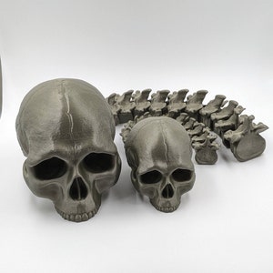 May include: A set of three 3D printed skulls, one large and two small, in a dark gray color. The skulls are arranged in a row, with the large skull in the center and the two smaller skulls on either side. The skulls are detailed and realistic, with visible teeth and eye sockets.