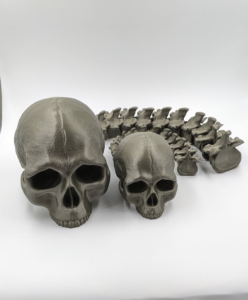 May include: A set of three 3D printed skulls, one large and two small, in a dark gray color. The skulls are arranged in a row, with the large skull in the center and the two smaller skulls on either side. The skulls are detailed and realistic, with visible teeth and eye sockets.