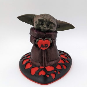 May include: A 3D printed figurine of a green alien character holding a red heart with the letters "AD" on it. The figurine is standing on a black heart-shaped base with red hearts cut out.