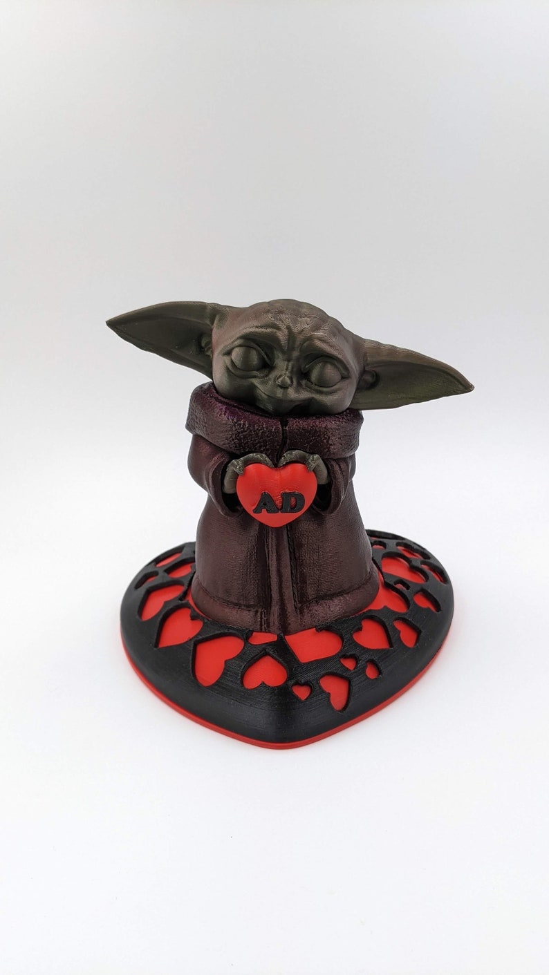 May include: A 3D printed figurine of a green alien character holding a red heart with the letters "AD" on it. The figurine is standing on a black heart-shaped base with red hearts cut out.
