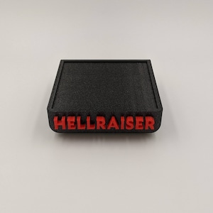 May include: A black, rectangular box with the word "HELLRAISER" in red, raised lettering. The box has a textured surface and rounded edges. The design is simple and bold.