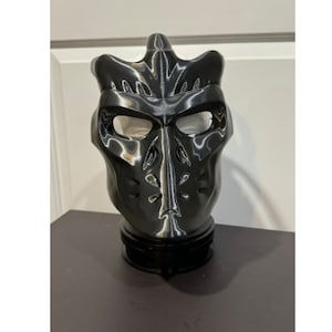 May include: A black plastic 3D printed mask with a detailed design. The mask has a pointed top and a wide, flat base.