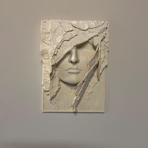 May include: A rectangular, off-white relief sculpture of a human face. The face is partially obscured by textured, cracked material, creating a dramatic effect. The sculpture is mounted on a light gray wall.