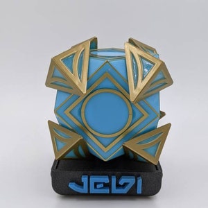 May include: A blue and gold geometric cube with a circular opening in the center, sitting on a black base with the word "JELVI" printed in blue.