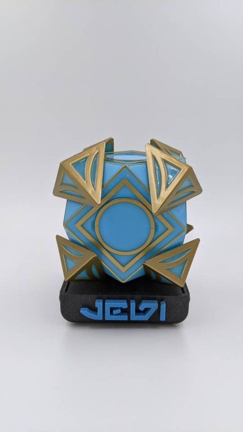 May include: A blue and gold geometric cube with a circular opening in the center, sitting on a black base with the word "JELVI" printed in blue.