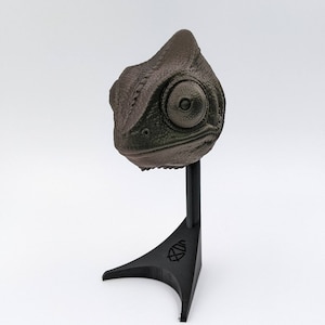 May include: A 3D printed chameleon head sculpture in a dark brown color, mounted on a black stand. The chameleon's eye is visible and has a green color.