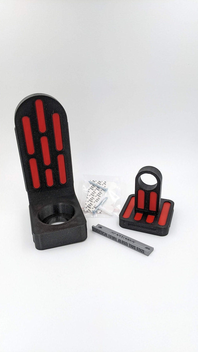 以下が含まれることがあります： 赤と黒のデザインの3Dプリントされた壁掛け。マウントには円形の開口部と長方形のベースがあります。マウントはライトセーバーを保持するように設計されています。画像には、「VERTICAL LEVEL ALONG THIS EDGE」というテキストが書かれた小さな紙も含まれています。