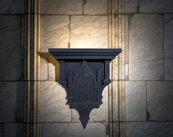 May include: A dark gray, gothic-style wall shelf with an arched window design. The shelf has a flat top and a decorative, pointed bottom. The background is a marble wall with vertical accents, creating a dramatic contrast.