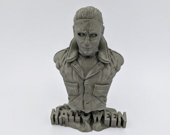 May include: A gray 3D printed bust of Michael Myers from the Halloween franchise. The bust is wearing a jacket and has a mask on. The bust is sitting on a base that says "HALLOWEEN".