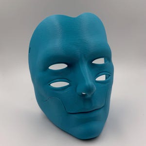 May include: A teal-colored mask with a human face design. The mask has eye and mouth openings, and a detailed texture. The mask is a solid color and appears to be made of a rigid material.