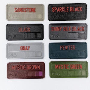 May include: Eight colour swatches labelled with the names "Sandstone", "Sparkle Black", "Black", "Shiny Silk Black", "Grey", "Pewter", "Mystic Brown", and "Mystic Green". Each swatch is made of PLA plastic.