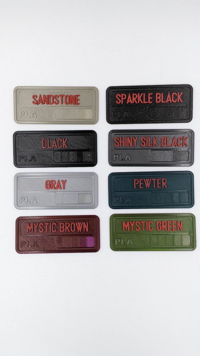 May include: Eight colour swatches labelled with the names "Sandstone", "Sparkle Black", "Black", "Shiny Silk Black", "Grey", "Pewter", "Mystic Brown", and "Mystic Green". Each swatch is made of PLA plastic.