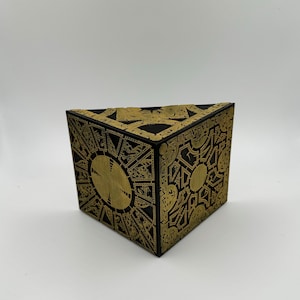 May include: A black and gold cube puzzle box with intricate designs on all sides. The box is made of wood and has a triangular shape.