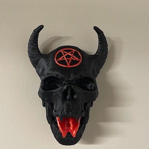 May include: A black skull with horns and a red pentagram on the forehead. The skull has a red tongue sticking out.