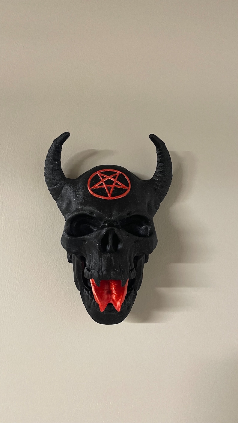 May include: A black skull with horns and a red pentagram on the forehead. The skull has a red tongue sticking out.