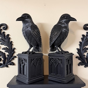 May include: Two black raven statues perched atop decorative pedestals, displayed on a matching black shelf. The ravens have detailed feather textures and stand in a gothic style. The shelf and pedestals feature arched designs.