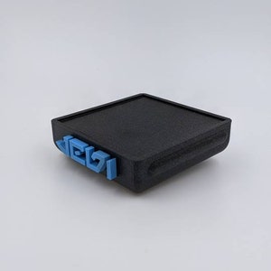 May include: A black rectangular box with a textured surface. The box has a blue inscription on the front that reads "LEGO".