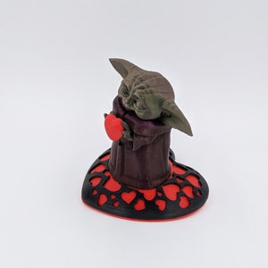 May include: A small green and brown figurine of a character holding a red heart. The figurine is standing on a black and red heart-shaped base.