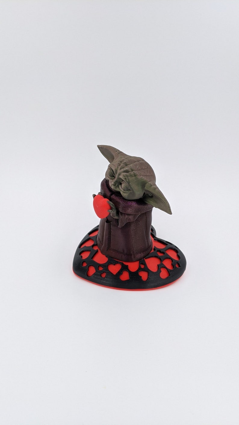 May include: A small green and brown figurine of a character holding a red heart. The figurine is standing on a black and red heart-shaped base.