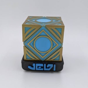 May include: A 3D printed cube with a geometric design in gold and blue. The cube is sitting on a black base with the word "JELSI" printed in blue.