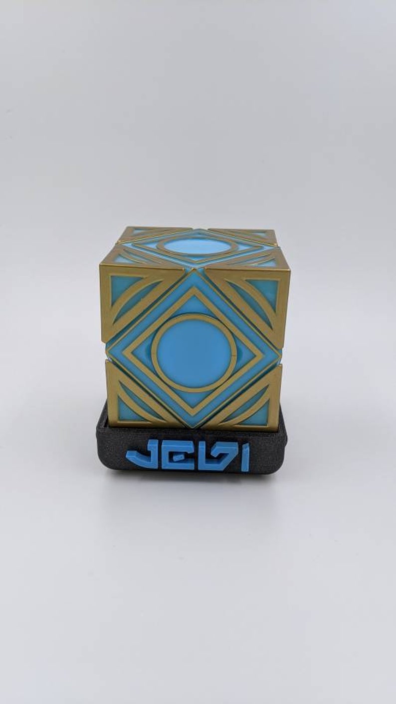 May include: A 3D printed cube with a geometric design in gold and blue. The cube is sitting on a black base with the word "JELSI" printed in blue.