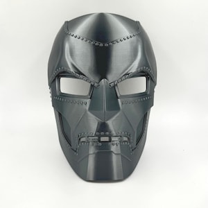 May include: A black plastic mask with a metallic finish and a detailed design. The mask has a pointed chin and a raised brow, with two eye holes and a small mouth opening. The mask is decorated with small, raised dots around the edges and on the forehead.