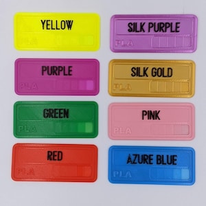 May include: Eight sample filament spools labelled with their colours and material type: yellow, silk purple, purple, silk gold, green, pink, red, and azure blue. Each spool is labelled "PLA".