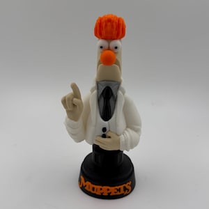 May include: A figurine of Dr. Bunsen Honeydew from The Muppets. The figure has an orange hairpiece, a white lab coat, and a black tie. The base is black with the word "Muppets" in orange.