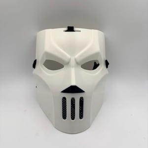 Puede incluir: Una máscara facial completa de color blanco con crestas angulares y agresivas en las cejas, aberturas para los ojos en forma de almendra, un pequeño detalle nasal triangular negro y tres ranuras de ventilación verticales en la barbilla.