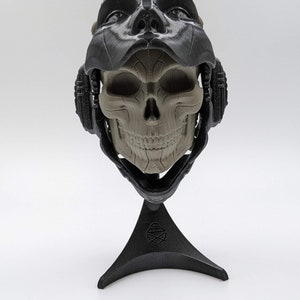 May include: A 3D printed skull sculpture with a black helmet and a stand. The skull has a detailed design and is painted in a light gray color.