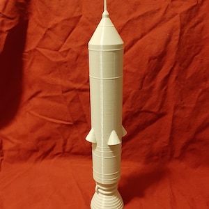 May include: A white 3D-printed rocket model. It has a conical top, a cylindrical body with horizontal bands, and triangular fins. The base is flared. The model is set against a red backdrop. The rocket is approximately 30 cm tall.