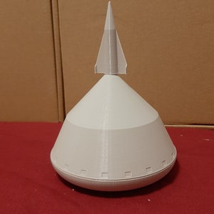 Op de afbeelding: Een wit 3D-geprint model van een raket met een puntige neuskegel en een ronde basis. De basis heeft een reeks kleine, gelijkmatig verdeelde cirkels rond de rand.