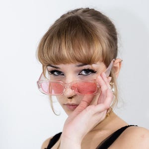 May include: A woman with blonde hair and bangs wearing pink sunglasses and a black lace top. She has a gold chain necklace with a cross pendant and a nose ring.