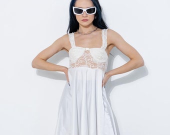 May include: A woman wearing a white satin slip dress with lace detailing, black furry leg warmers, and white sunglasses.
