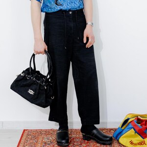May include: A person wearing a blue short-sleeved shirt with a black dragon print, black pants, and black clogs. They are holding a black handbag. There are two colorful bags in the background.