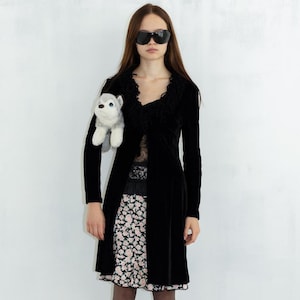 May include: A woman wearing a black velvet jacket with a lace trim, a floral skirt, black tights, and black heels. She is holding a small stuffed animal.