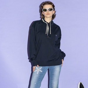 May include: A person wearing a dark blue long-sleeved shirt with a zipper and a white flower design on their jeans. They are wearing sunglasses and standing in front of a purple background.