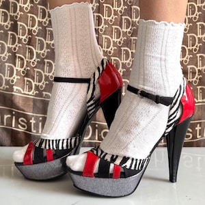 May include: Platform high heels with a zebra print, red and black accents. The heels have a black ankle strap and a silver platform sole. The shoes are paired with white socks.