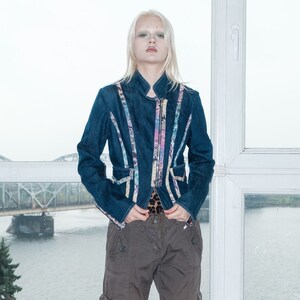 May include: A woman wearing a blue denim jacket with colourful trim and brown cargo trousers. She is standing in front of a window with a view of a bridge and water.