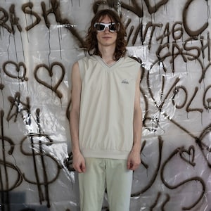 May include: A person wearing a beige sleeveless shirt with a small logo on the chest, light green pants, and neon yellow sneakers. The person is standing in front of a wall covered in black spray paint.