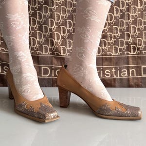May include: Tan leather high-heeled shoes with a square toe and a snakeskin-like design on the toe box. The heels are a dark wood color. The shoes are worn with white patterned stockings.