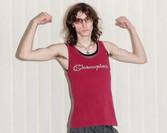 May include: A person wearing a red tank top with the word "Champion" printed on it, black cargo trousers, and brown sandals. The person is flexing their biceps.