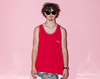 May include: A man wearing a red tank top with a white logo, camouflage shorts, and black boots. The tank top has a white trim around the neck and armholes.