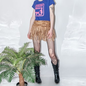 Puede incluir: Una mujer con una camiseta azul con el texto "CHICKS RULE" en letras rosas y una falda marrón con flecos. Lleva medias de rejilla negras y botas negras.