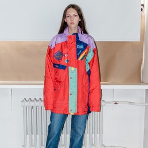 May include: A red, blue, green and purple ski jacket with various patches and logos. The jacket has a zip closure and a hood. The model is wearing blue jeans and brown trainers.