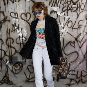 May include: A person wearing a black faux fur coat, white jeans, and orange pointed-toe heels. They are also wearing blue sunglasses and a white graphic tee. A leopard print handbag is held in their hand.