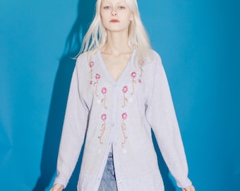 May include: A light blue cardigan with a V-neck and long sleeves. The cardigan features embroidered pink and white flowers. The model is wearing a denim skirt. A neon sign that says "BAR" is in the lower left corner.